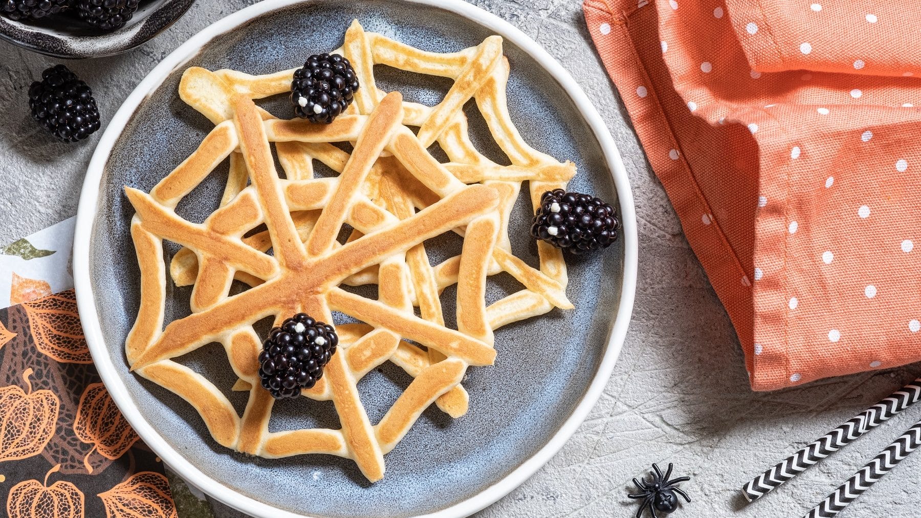 Zwei Spinnennetz-Pfannkuchen mit Brombeeren auf einem gemusterten Teller auf einem grauen Tisch. Um den Teller herum liegen Servietten, ein geschirrtuch, eine Schale mit Brombeeren und eine Plastik-Spinne. Außerdem liegen am rechten Bildrand zwei gemusterte Strohhalme.