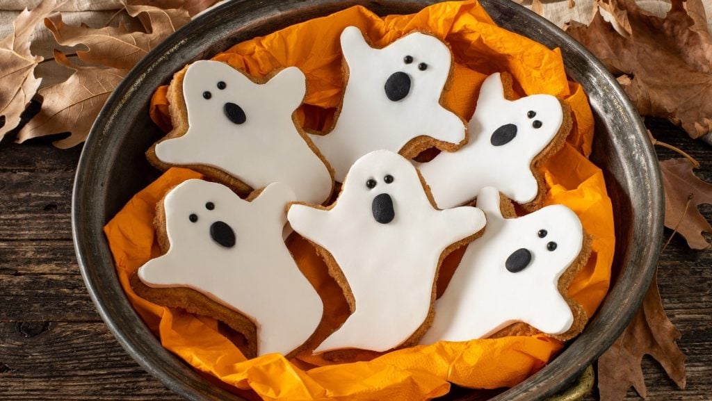 Sechs Geister-Kekse mit Fondant liegen in einer Schale mit orangener Serviette. Die Schüssel steht auf einem braunen Holztisch. Um die Schüssel herum liegen braune Laubblätter.