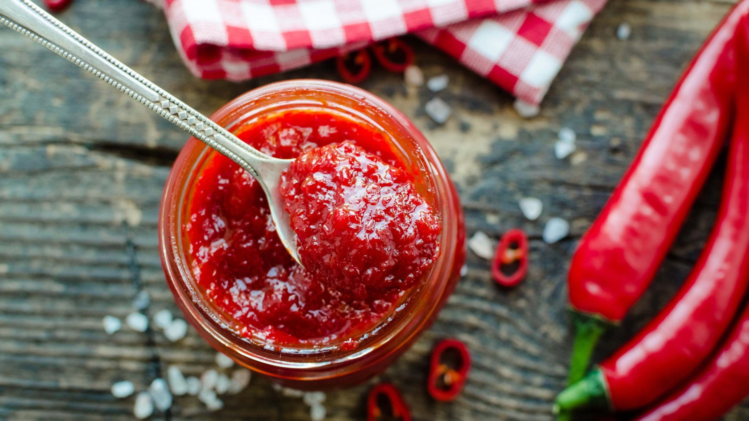 Draufsicht von oben: Ein Glas mit einer Tomaten-Chili-Marmelade steht auf einem Tisch. Daneben liegen zwei Chilischoten.. Die Marmelade heißt Red Hot Chili Jam..