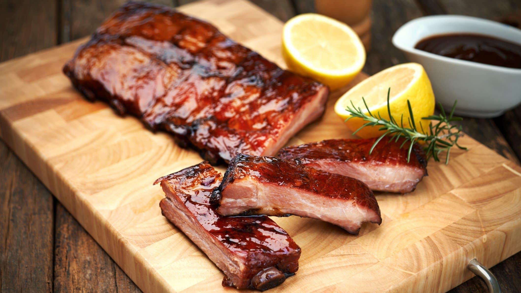 Ofen Spareribs auf Holzbrett mit Dillzweig und Zitronenhälften. Daneben Schale mit Sauce. Angerichtet auf dunklem Holztisch. Frontalaufnahme.