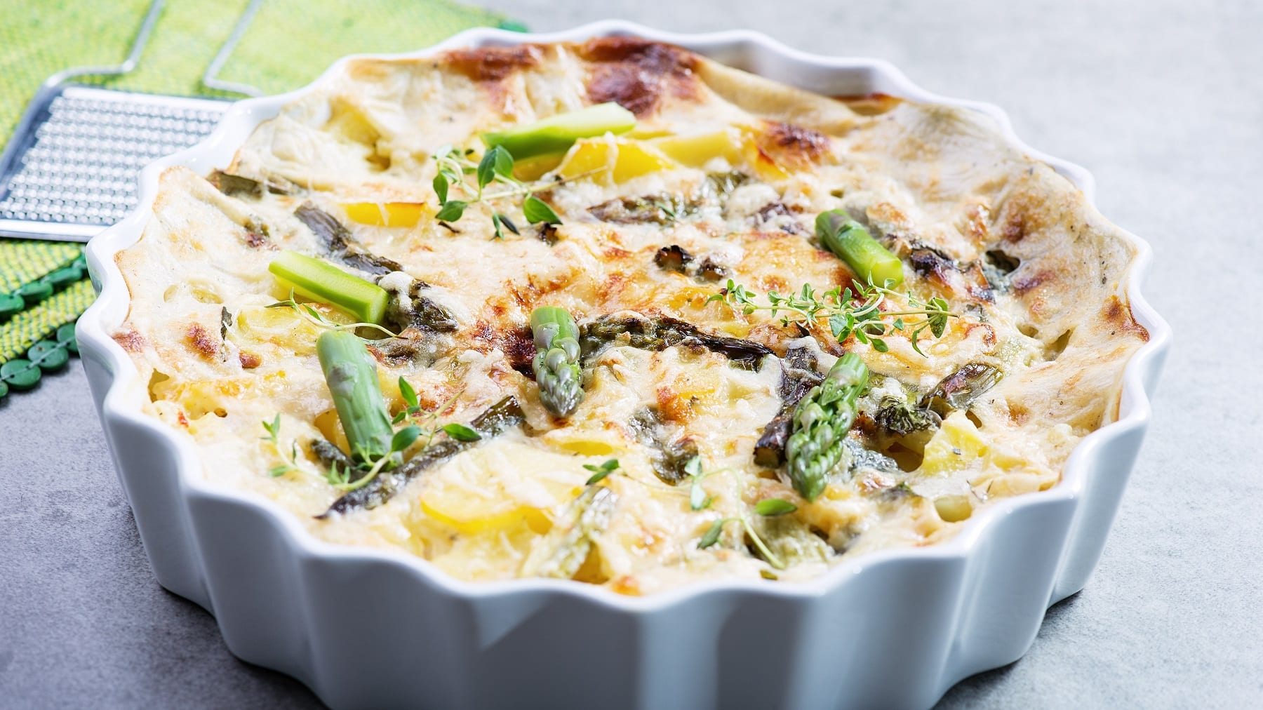 Eine weiße Auflaufform mit Kartoffel-Spargelgratin auf einem grauen Tisch. Hinten links unscharf ein grünes Tuch mit einer Metallreibe zu erkennen.