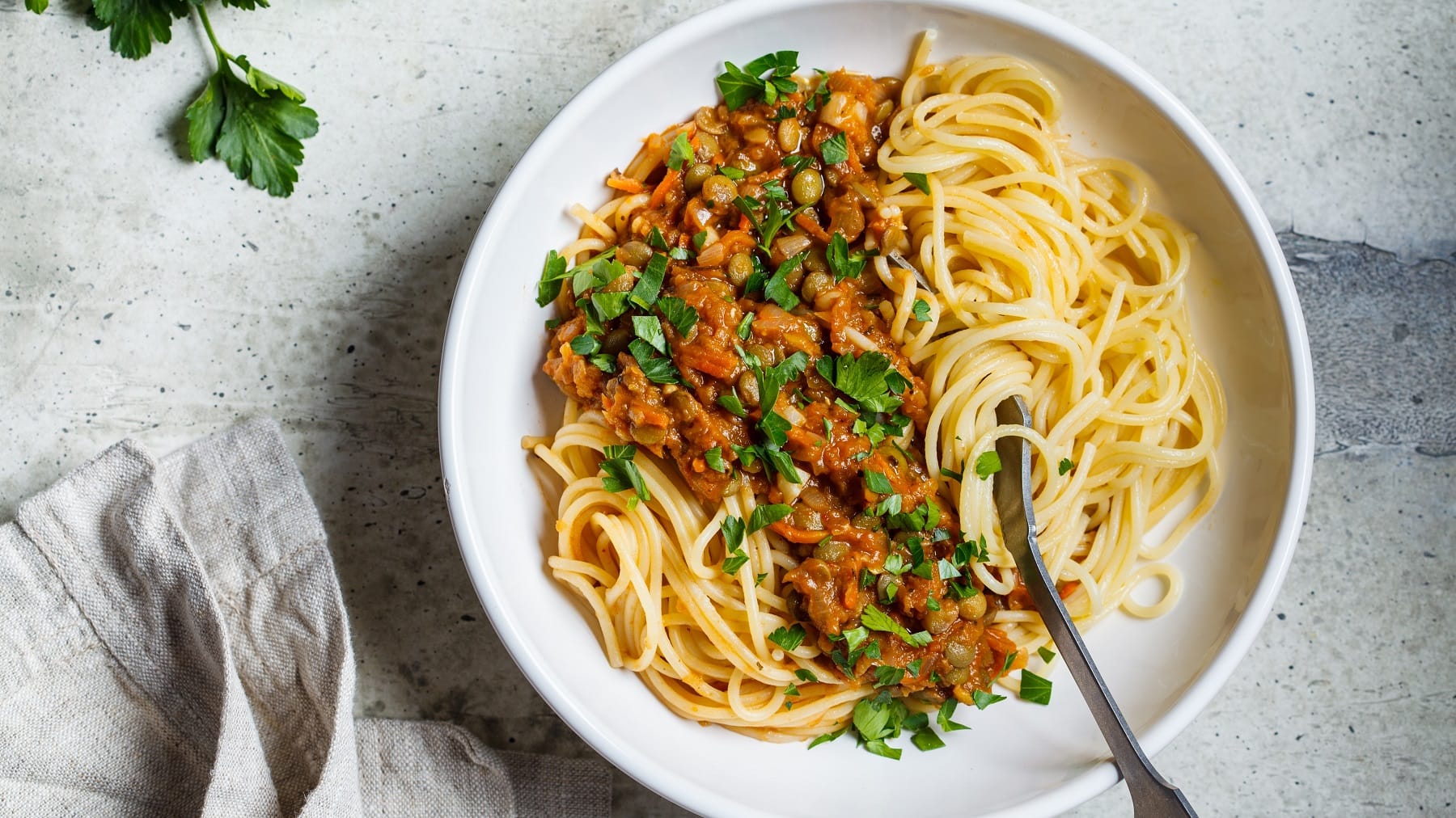 Ein weißer Teller mit Spaghetti und Linsen-Bolognese auf einem grauen Steintisch direkt von Oben fotografiert. Links vom Bild außerdem, Petersilie und ein graues Tuch. Rechts im Bild eine silberne Gabel, die in den Teller ragt.