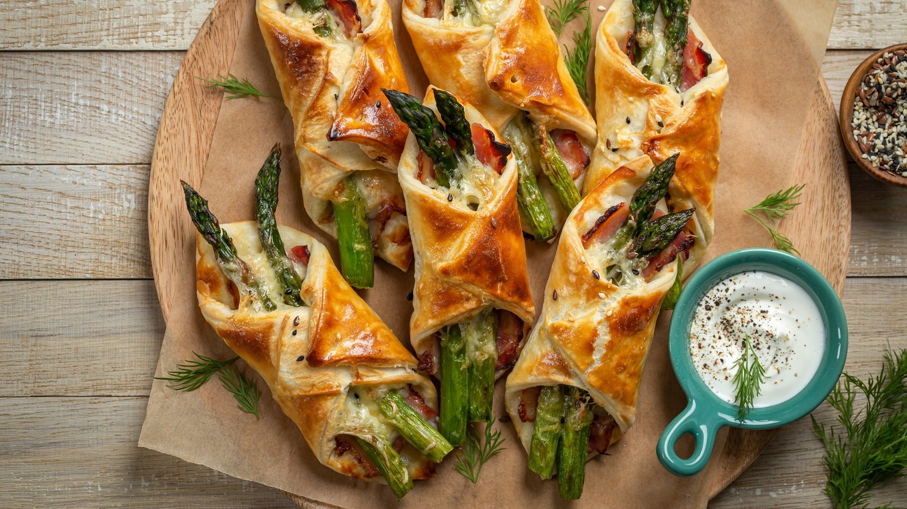 Blätterteigtaschen mit grünem Spargel und Bacon auf einem runden Holzbrett. Daneben ein weißer Dip.