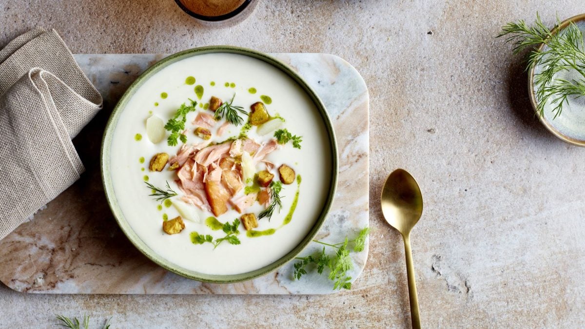 Spargelcreme mit Brezel und Lachs in einer Schüssel auf einem marmoriertem Brett vor beigem Hintergrund. Rechts ein goldener Löffel und links ein beiges Geschirrtuch.